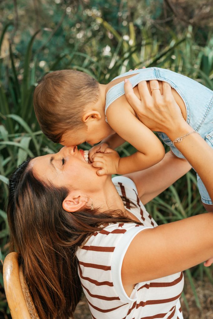 Una madre sonriente sostiene a su hijo pequeño, ambos riendo y jugando en un ambiente natural con vegetación de fondo.